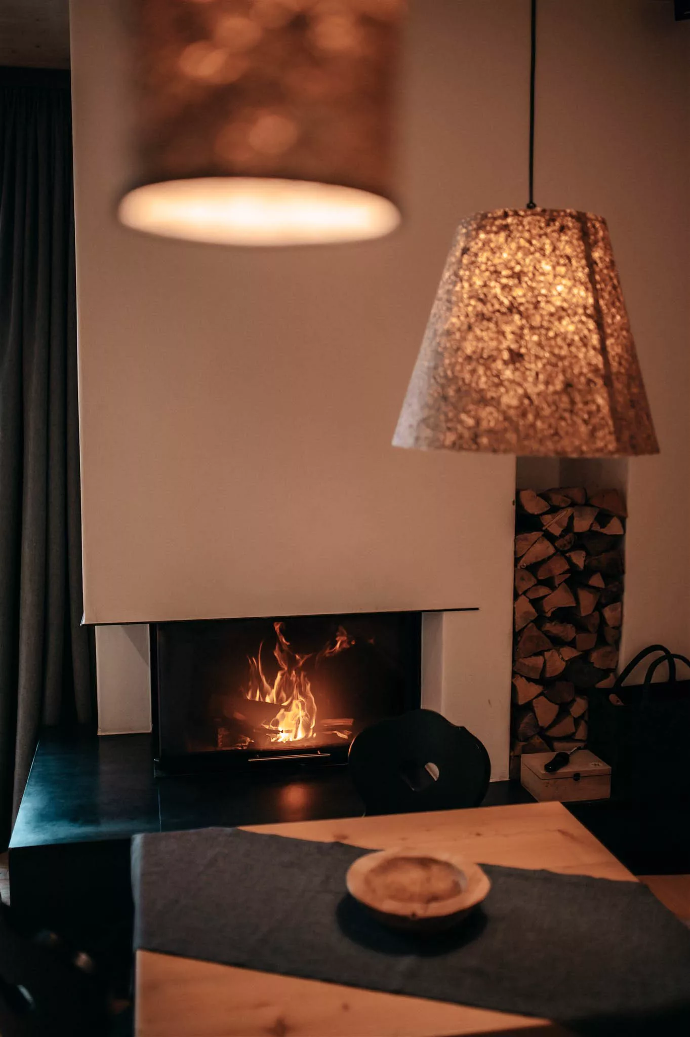 Ein gemütlicher Raum mit einem brennenden Kamin, daneben gestapeltem Brennholz und zwei Hängelampen im Vordergrund. Davor steht ein Holztisch mit einer flachen Schale. Warme, einladende Atmosphäre.