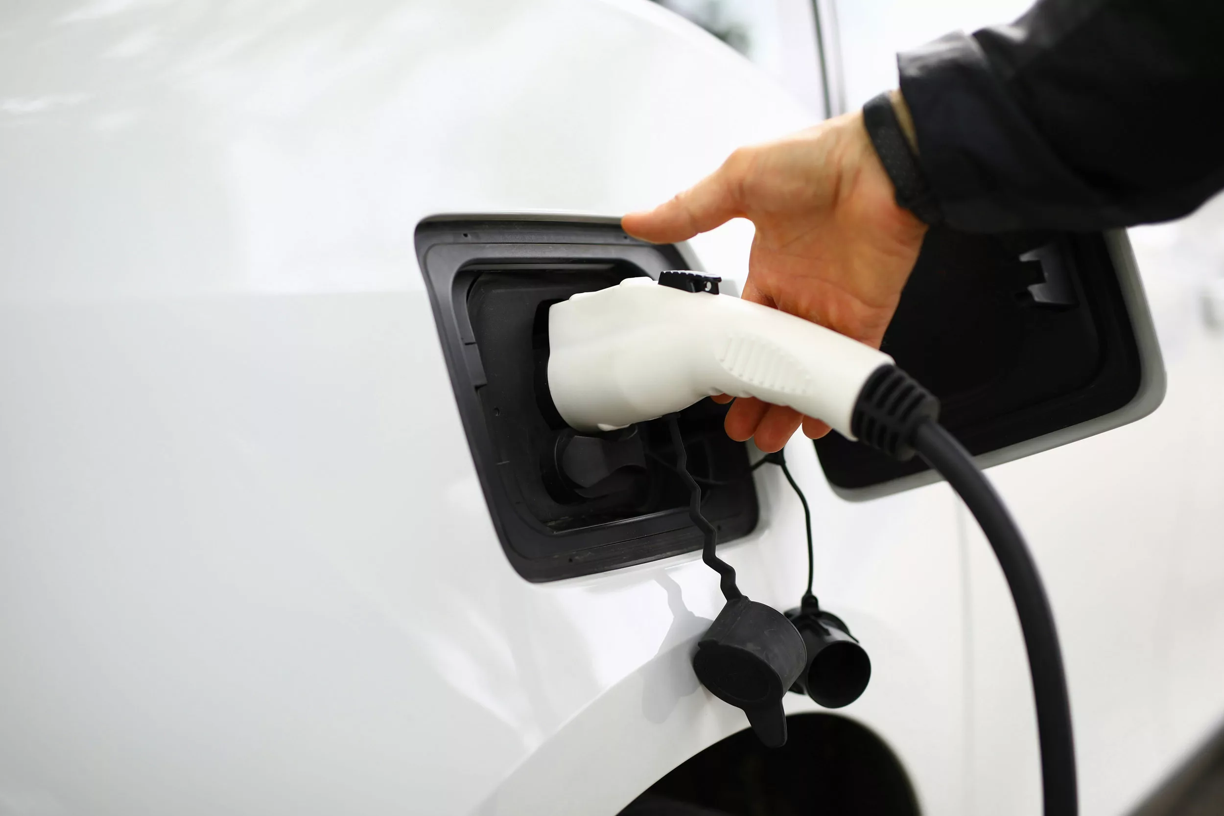 Eine Person steckt ein Ladekabel in den Anschluss eines weißen Elektrofahrzeugs, das im Freien geparkt ist.
