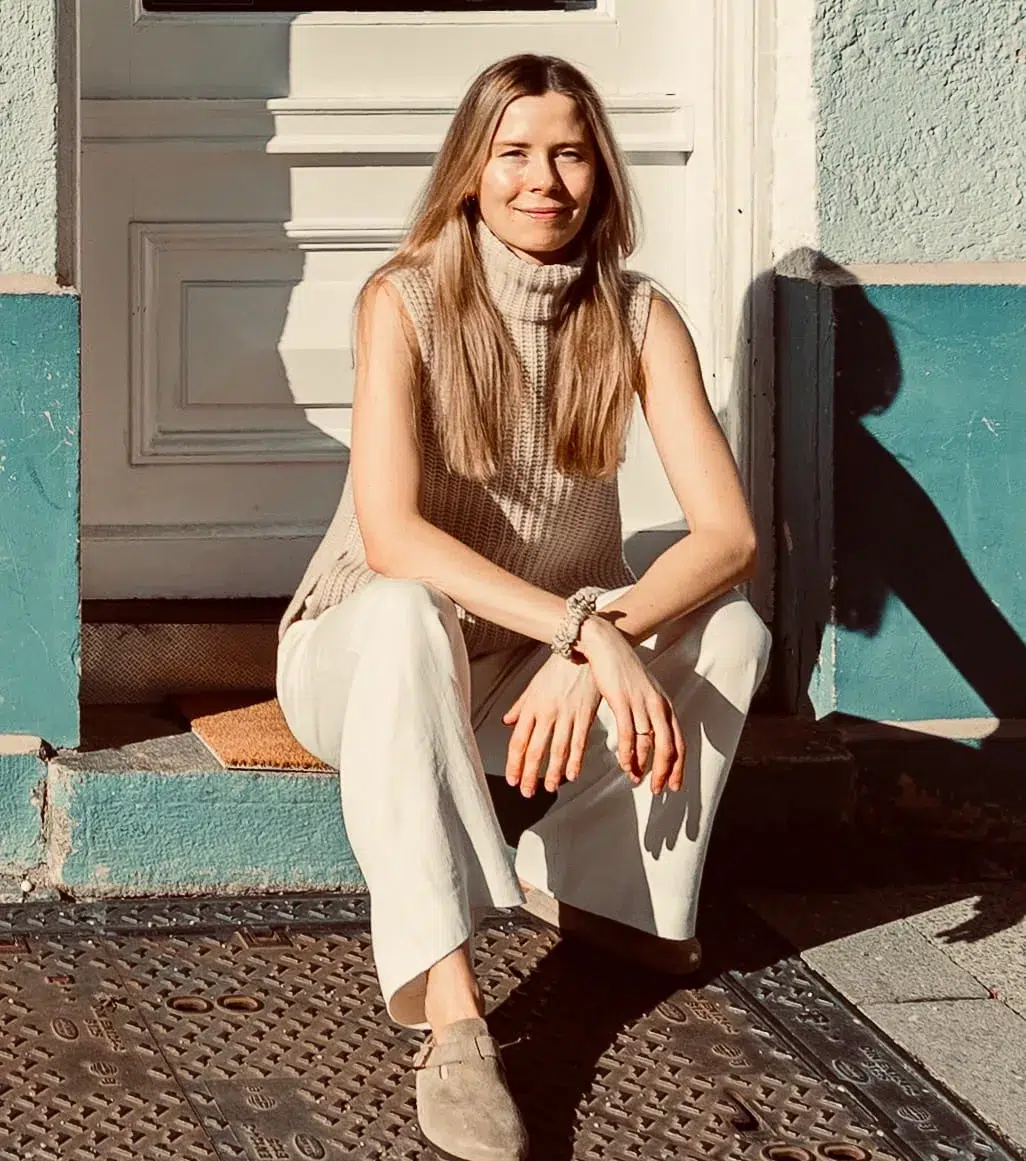 Eine Frau mit langen Haaren, die einen ärmellosen Rollkragenpullover und eine weiße Hose trägt, sitzt im Sonnenlicht auf einer Türschwelle und lächelt in die Kamera.