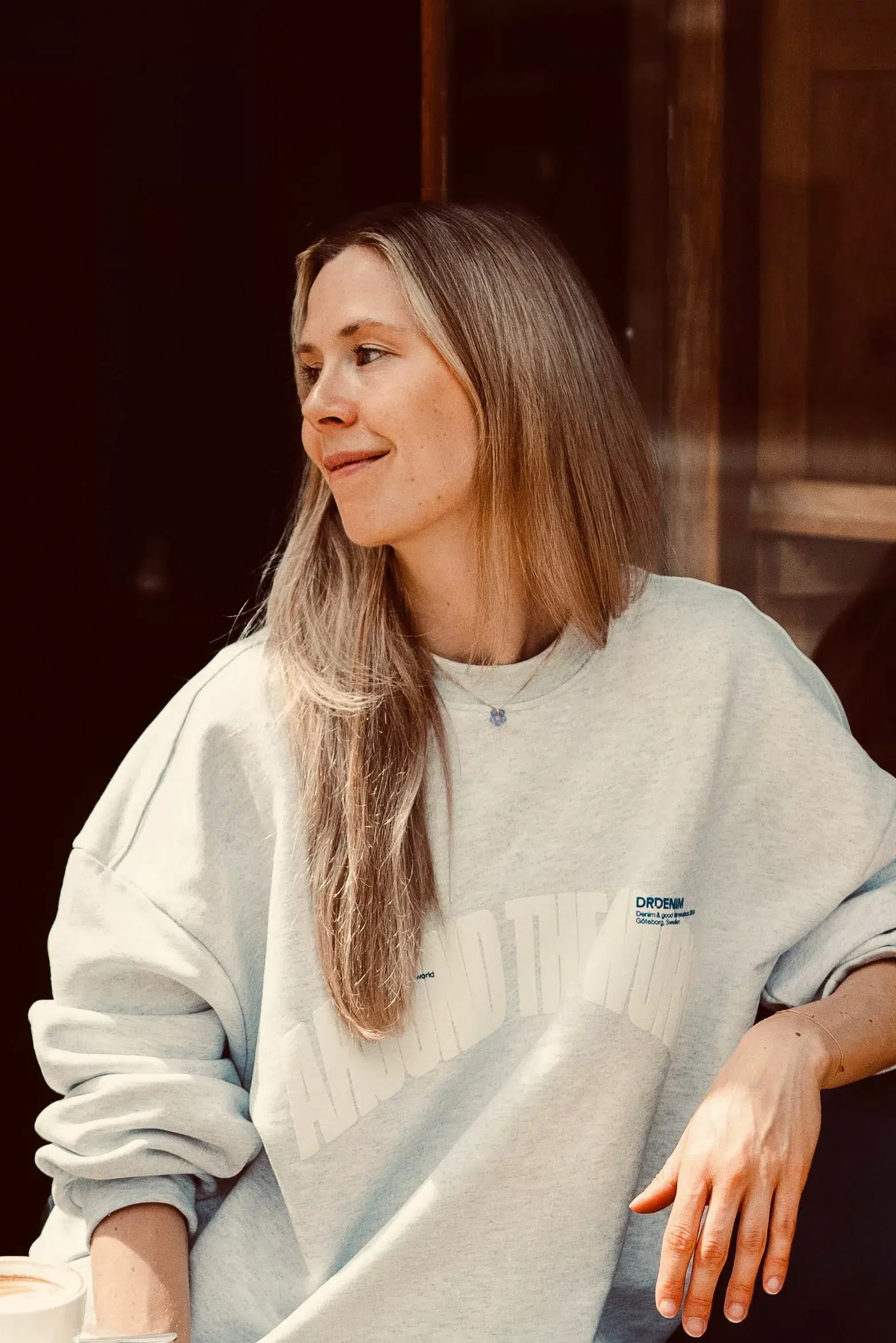 Eine Frau mit langen blonden Haaren sitzt an einem Tisch, lächelt und schaut zur Seite. Sie trägt ein hellgraues Sweatshirt und hat einen Arm neben einer Kaffeetasse auf dem Tisch abgelegt.